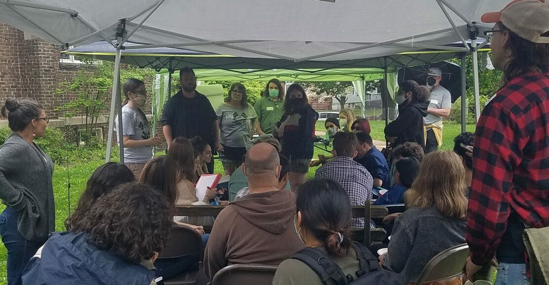A photo of our two facilitators leading our workshop outside under a tent on the grass with many people seated and standing around them listening.