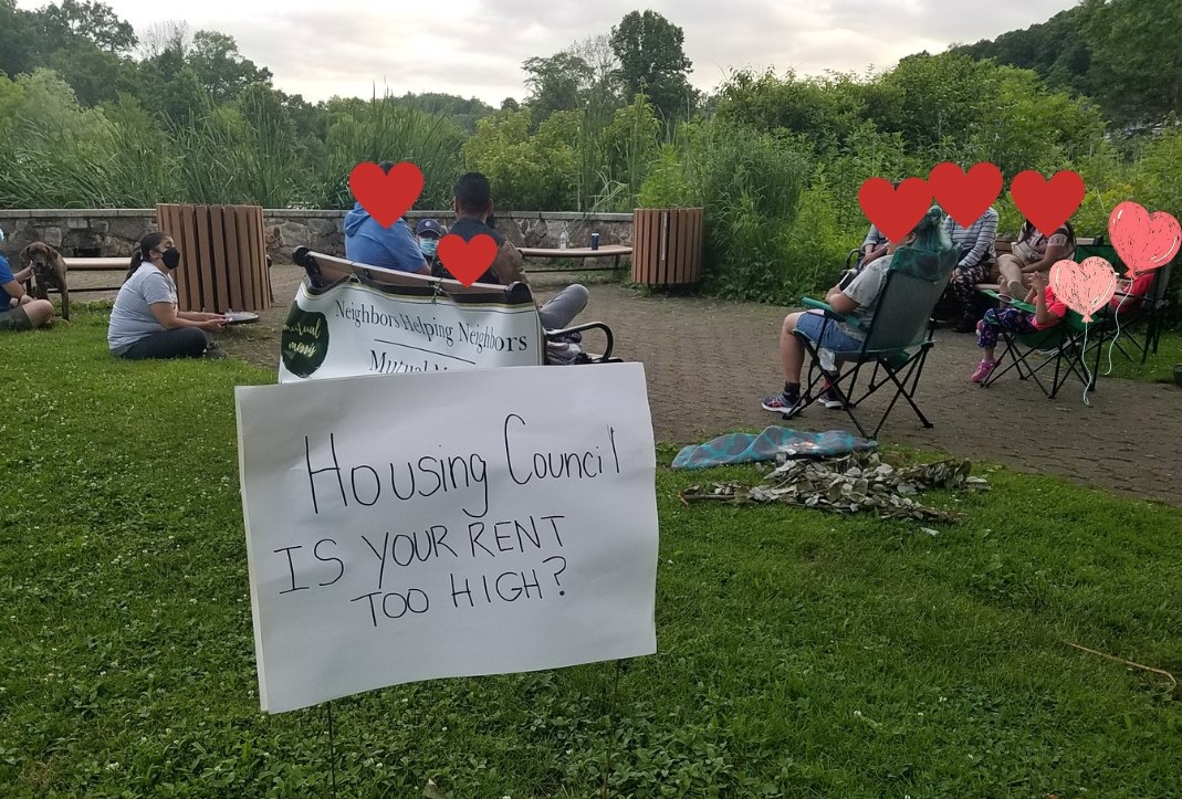 A photo of a group of people sitting in a park with a hand-drawn sign in the foreground that reads "Housing Council. Is your rent too high?" and graphics of red and pink hearts photoshopped over to cover individuals' faces for privacy.